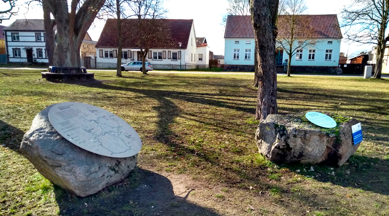 Ein Dorfplatz mit Bäumen und großen Steinen mit Schildern.