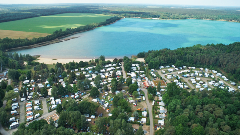 PdM 05-25 Start neu Bild enthält einen See, umgeben von Wald und Feldern und einen Campingplatz.