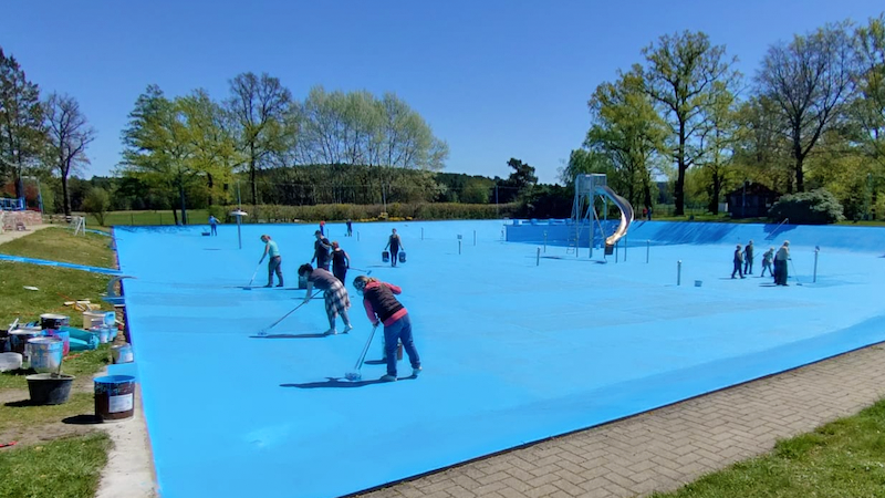 PdM 06-25 top Bild enthält zahlreiche Menschen, die blaue Farbe auf dem Boden eines Schwimmbeckens verteilen.