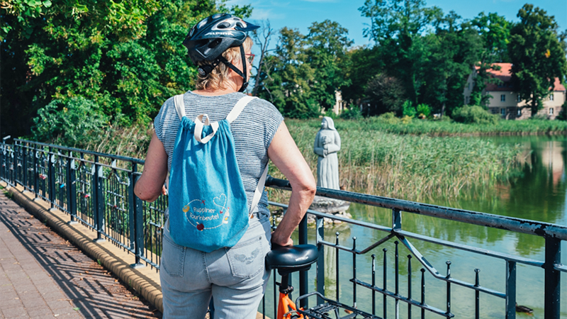 Bild enthält eine Frau mit Fahrrad auf einer Brücke, die einen Rucksack mit der Aufschrift „Ruppiner Tour´nbeutel“ trägt.